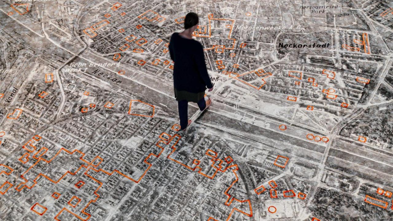 Eine Person läuft über eine auf einen Teppich applizierte Bodengrafik, die die zerbombte Stadt Mannheim von oben zeigt.