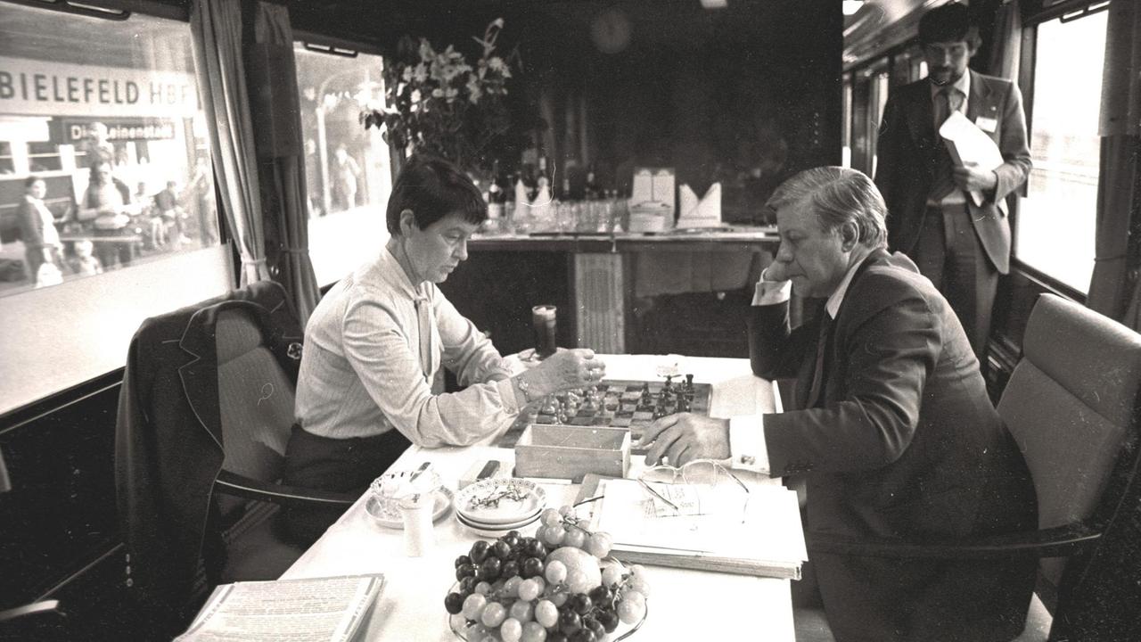 Helmut und Loki Schmidt spielen Schach in einem Eisenbahnzug, auf dem Tisch steht eine Obstschale, im Hintergrund nähert sich ein Referent mit Papieren