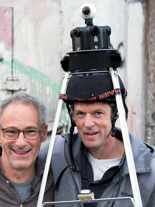 Regisseur Dani Levy und Kameramann Filip Zumbrunn am Drehort der Episode "Liebe/Love" vor der Sperrmauer. Zumbrunn hat ein auffälliges Gerüst auf dem Kopf, in dem sich seine Kamera befindet. Beide lachen in die Kamera. Zumbrunn umarmt Levy.