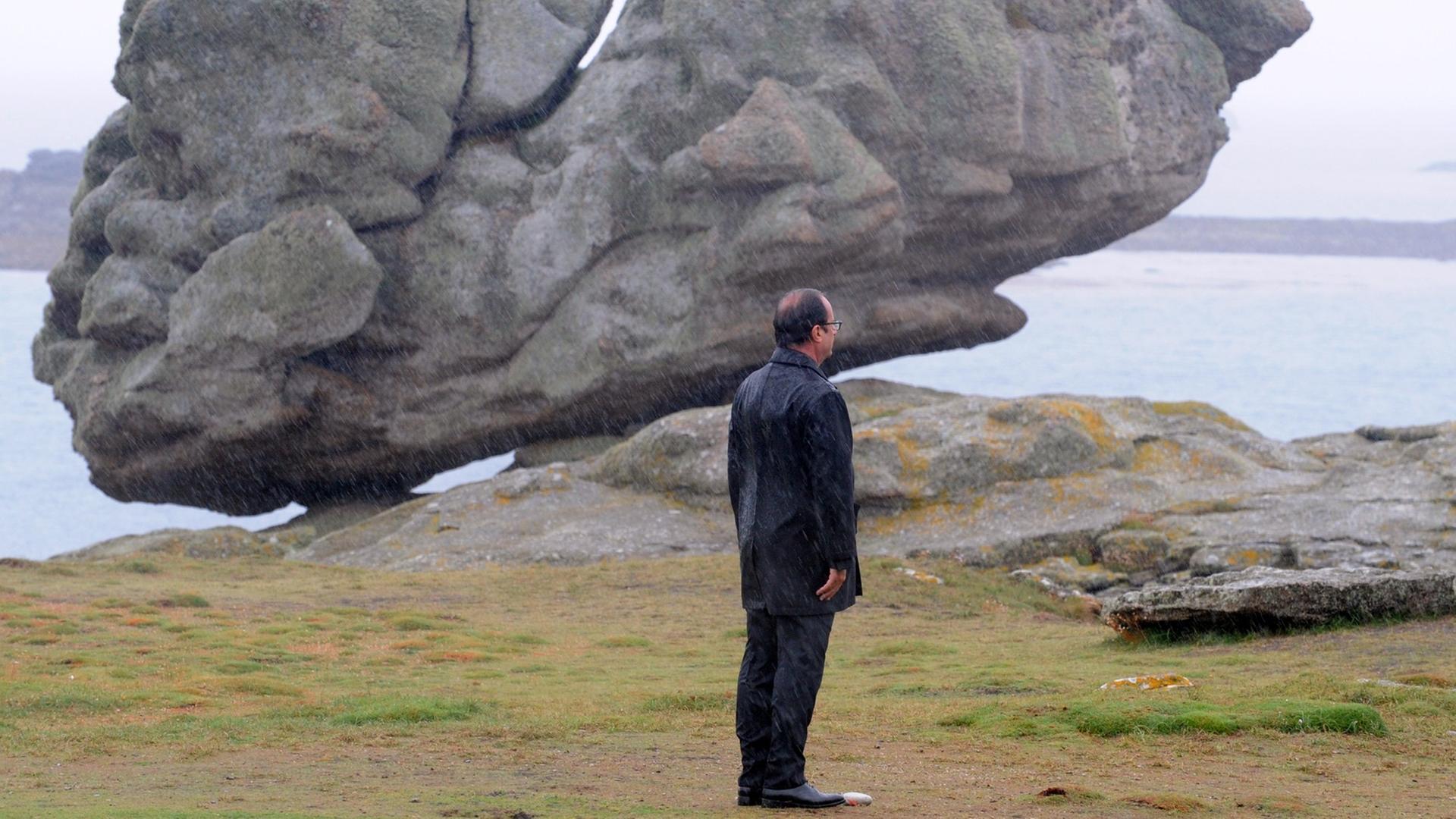 Francois Hollande steht an der Küste vor einem großen Felsen im Regen, man sieht ihn von hinten.