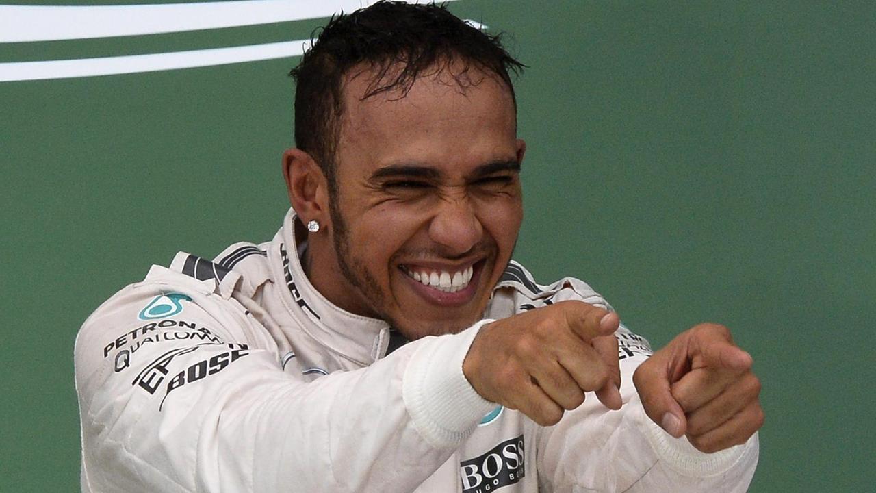 Hamilton im weißen Rennfahrer-Dress. Er zeigt mit beiden Händen lachend auf jemanden.