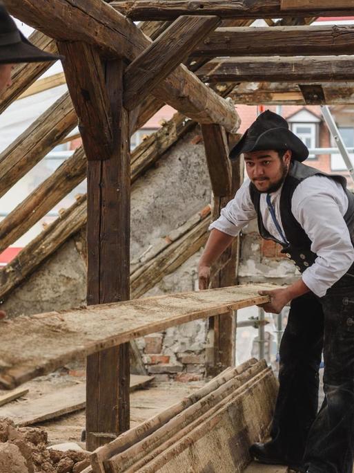 Zwei Zimmermannsgesellen, die auf Wanderschaft sind, arbeiten auf einer Baustelle in Bamberg. Sie halten gemeinsam ein Holzbrett.