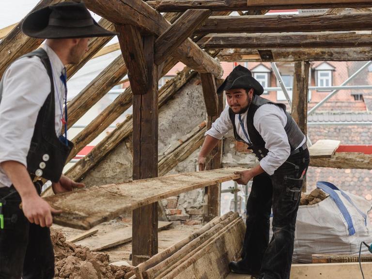 Zwei Zimmermannsgesellen, die auf Wanderschaft sind, arbeiten auf einer Baustelle in Bamberg. Sie halten gemeinsam ein Holzbrett.