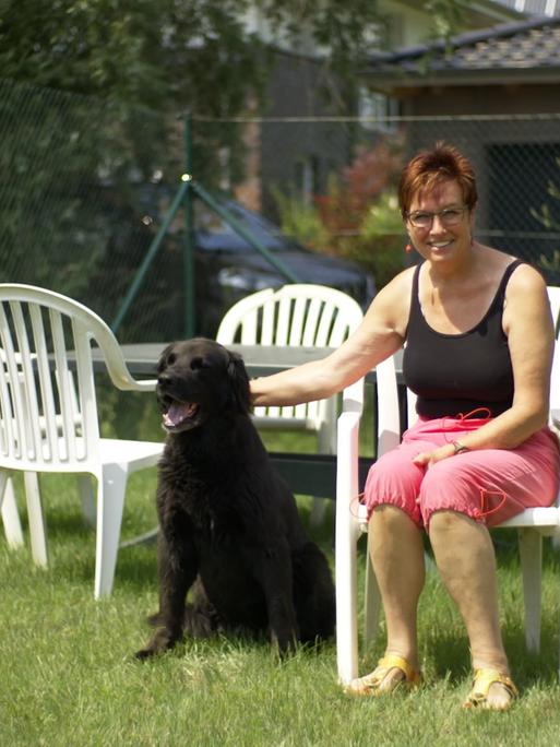 Szene aus dem Dokumentarfilm "Monobloc": Eine Frau sitzt im Garten auf einem weißen Plastikstuhl, um sie herum stehen weitere dieser Stühle. Die Frau streichelt einen Hund, der neben ihr auf dem Rasen sitzt.
