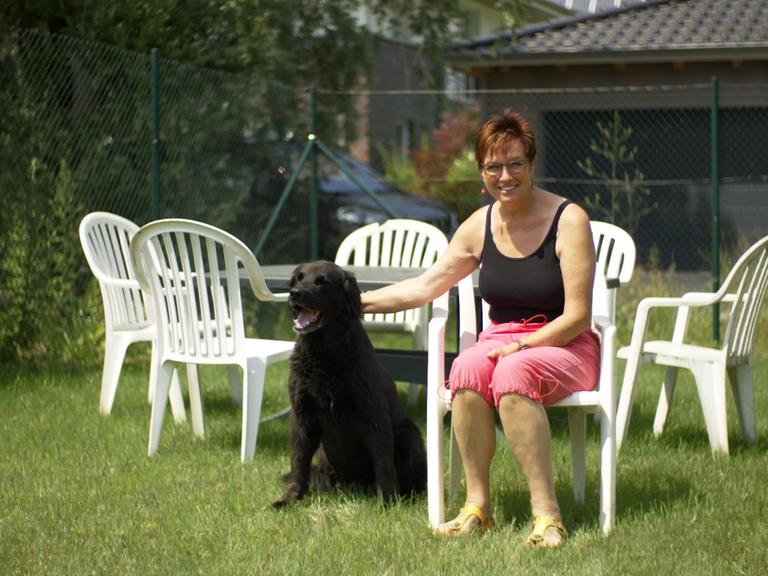 Szene aus dem Dokumentarfilm "Monobloc": Eine Frau sitzt im Garten auf einem weißen Plastikstuhl, um sie herum stehen weitere dieser Stühle. Die Frau streichelt einen Hund, der neben ihr auf dem Rasen sitzt.