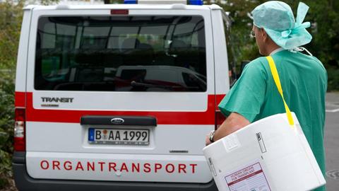 Der Bote trägt grüne Hosen, einen grünen Kittel und eine Haube. Er bringt den weißen Behälter, der über seine Schulter hängt, zu einem Transporter mit der Aufschrift "Organtransport".