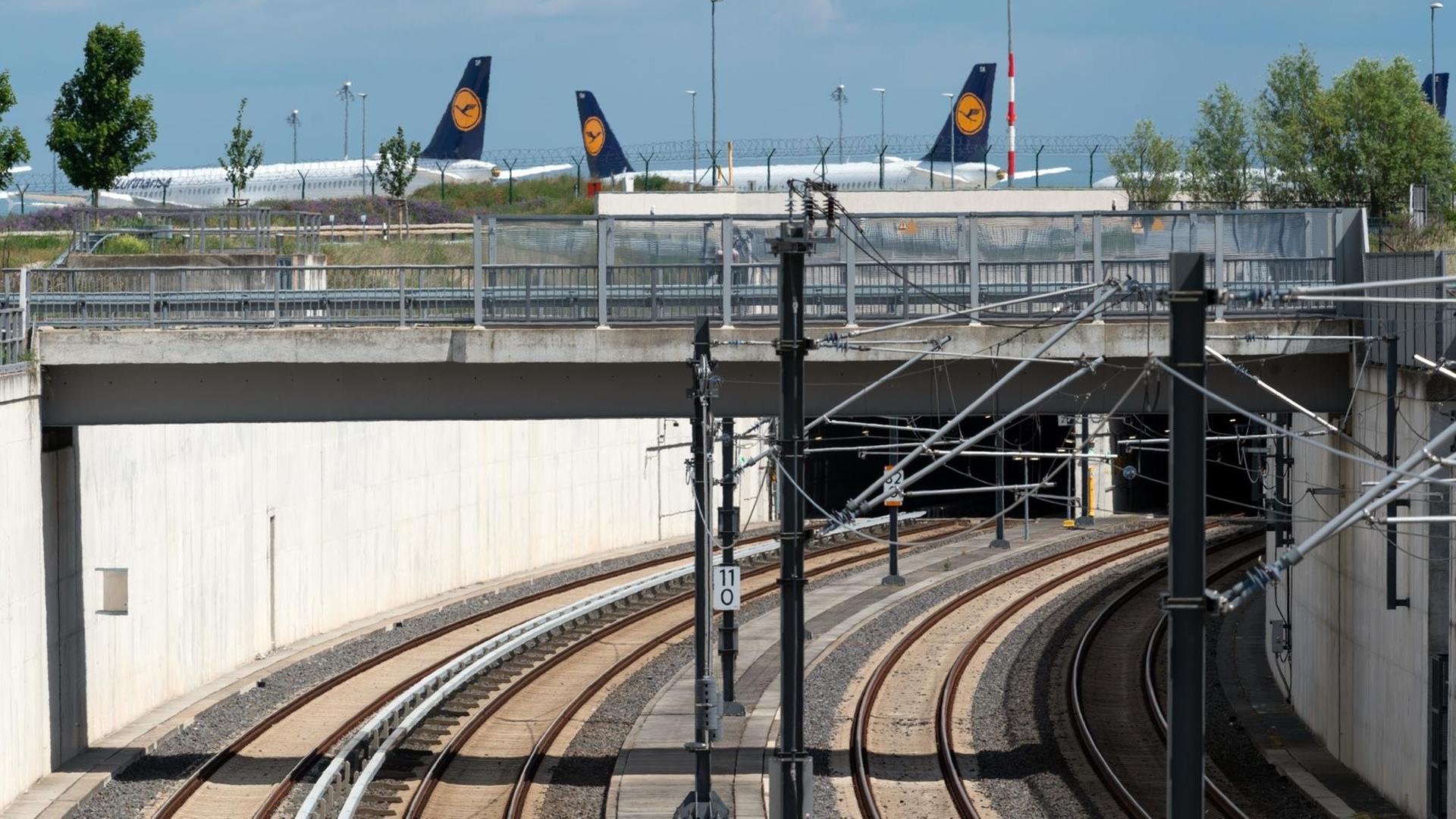 02.06.2020, Brandenburg, Potsdam: Die Schienen der S-Bahn (l) und der Regionalbahn führen in einen Tunnel zum unterírdischen Bahnhof des Hauptstadtflughafens. Im Hintergrund stehen abseits des Rollfeldes Flugzeuge der Fluggesellschaft Lufthansa. Foto: Soeren Stache/dpa-Zentralbild/ZB | Verwendung weltweit