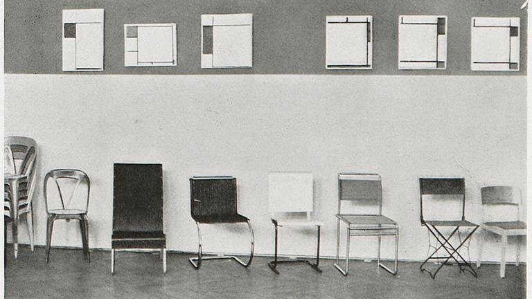 Ausstellung in Frankfurt im Jahre 1929 "Der Stuhl" Schwarzweiß Fotografie zeigt Stühle aufgereiht vor Gemälden von Mondrian.