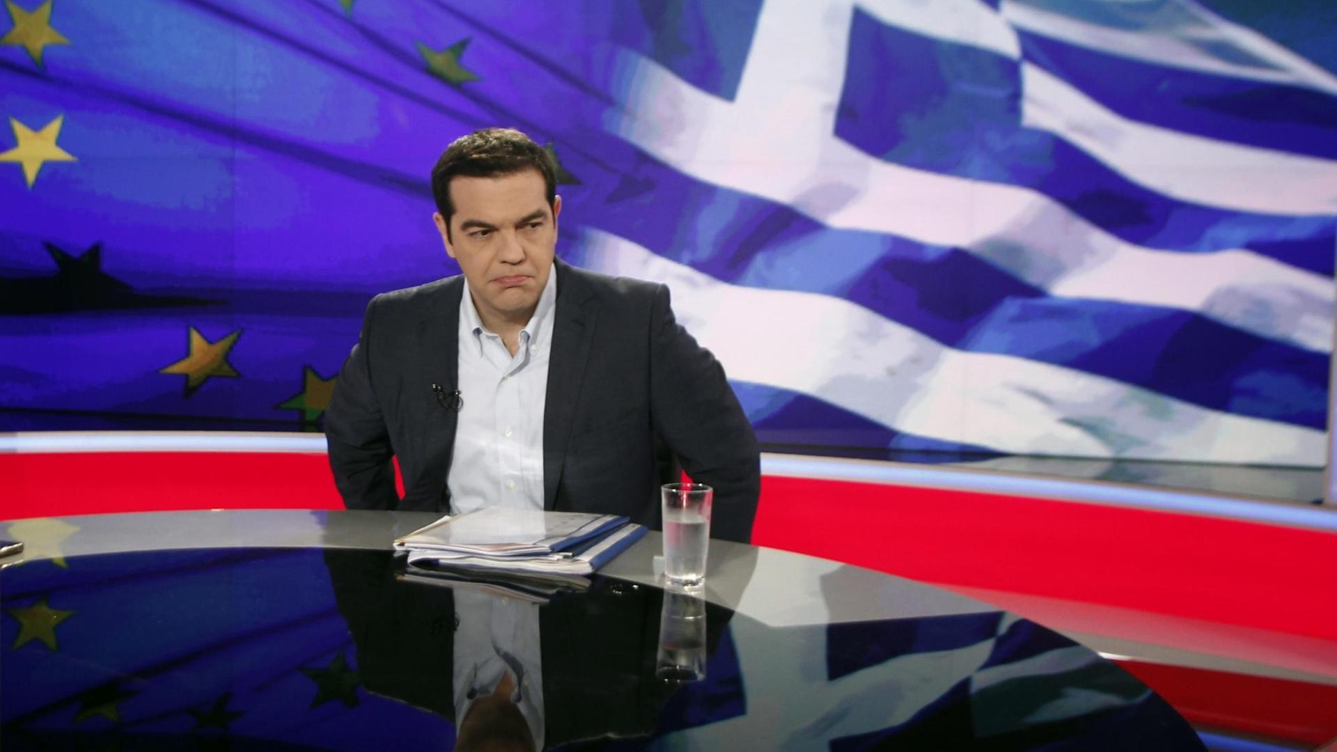 Tsipras sitzt vor einem Bild der griechischen und der europäischen Fahne.
