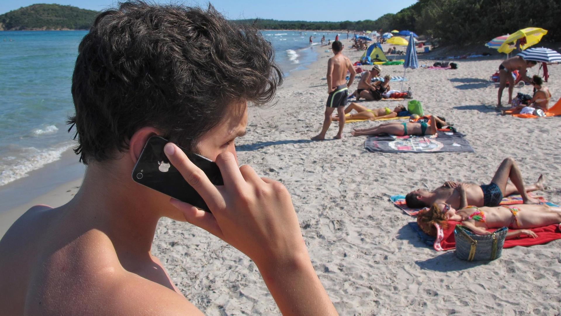 Ein junger Mann telefoniert an einem Sandstrand, im Hintergrund Sonnenbadende und das Meer.