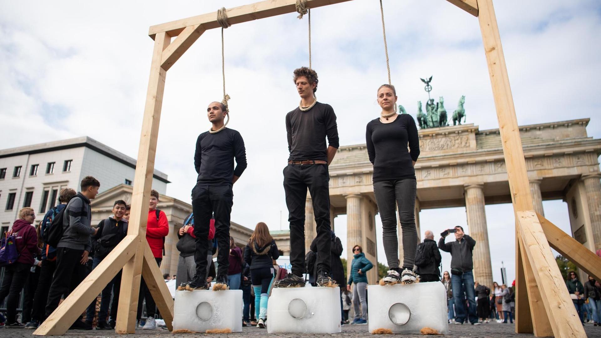 Drei Demonstranten stehen unter einem Galgen mit einem Seil um den Hals auf Eisblöcken. Dahinter ist das Brandenburger Tor.