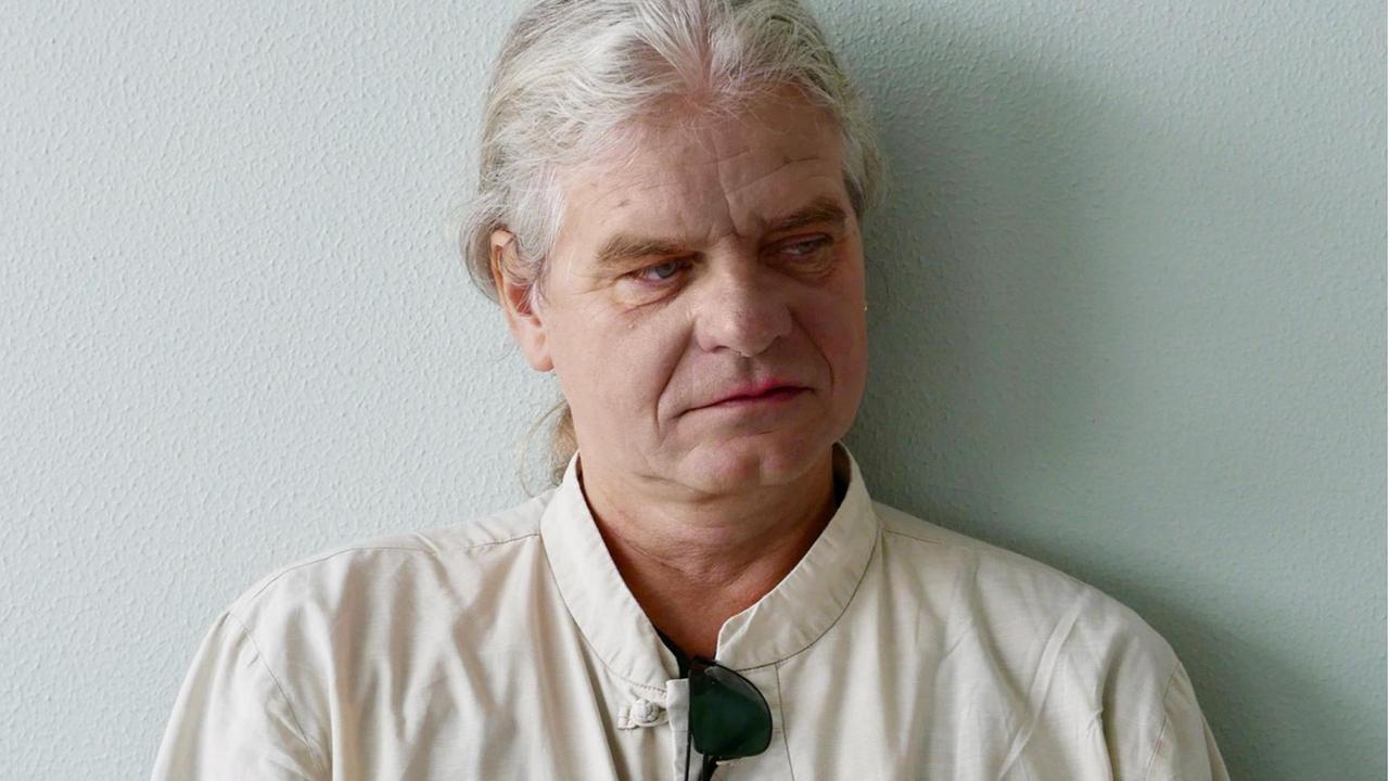 Der Religionswissenschaftler Volker Küster trägt sein weißes Haar im Nacken zu einem Zopf gebunden, im Kragen seines weißen Hemdes, steckt eine dunkle Sonnenbrille, sein Blick ist nachdenklich zur Seite gewandt.