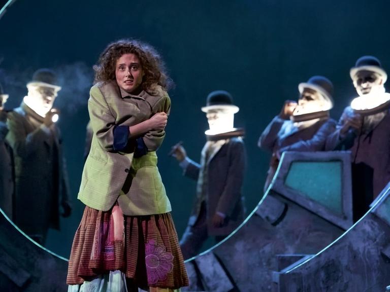 Szene aus der Inszenierung des Musiktheaters "Momo" in München mit der Hauptfigur Momo im Vordergrund und den grauen Männern im Hintergrund