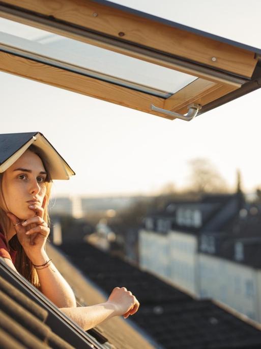 Eine junge Frau schaut nachdenklich aus einem Dachfenster mit einem aufgeschlagenen Buch über dem Kopf.