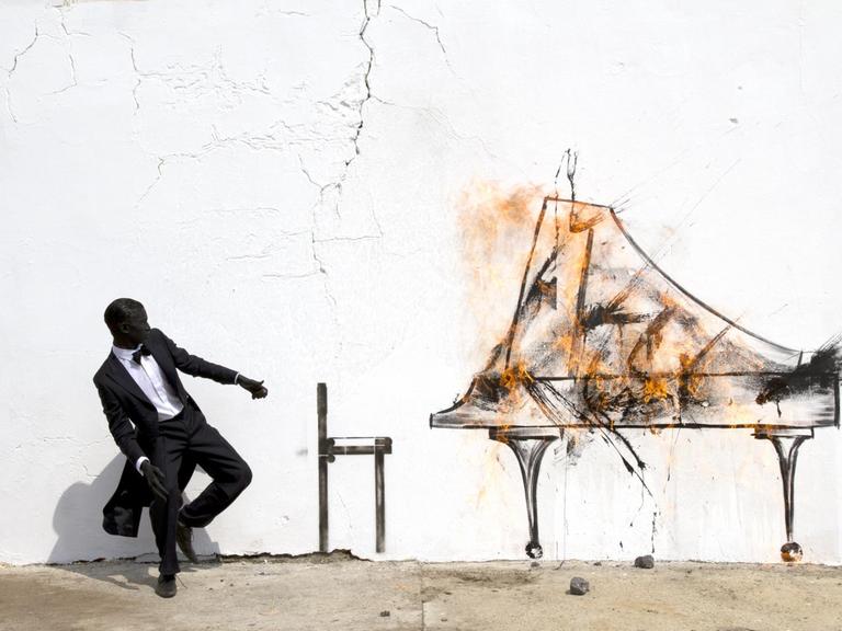Auf dem Bild "PIANO CHAIR" des südafrikanischen Streetart-Künstlers Robin Rhode sind an eine weiße Mauer ein Klavierstuhl und ein brennender, geöffneter Flügel gemalt. Im rechten Teil des Bildes hinter dem Klavierstuhl steht ein im Frack des Konzertpianisten gekleideter Mann, der in einer vor dem Feuer zurückschreckenden Pose festgehalten wurde.