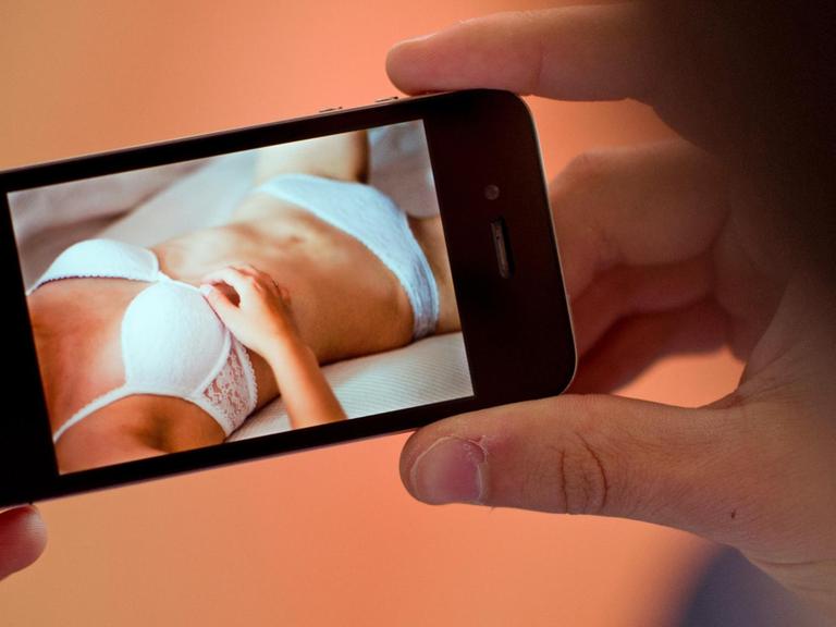 Ein Mann hält ein Smartphone in der Hand, auf dem ein erotisches Foto einer jungen Frau zu sehen ist.
