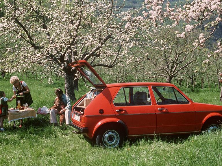 Eine Familie ist mit ihrem roten VW-Golf ins Grüne gefahren und veranstaltet ein Picknick unter blühenden Obstbäumen.