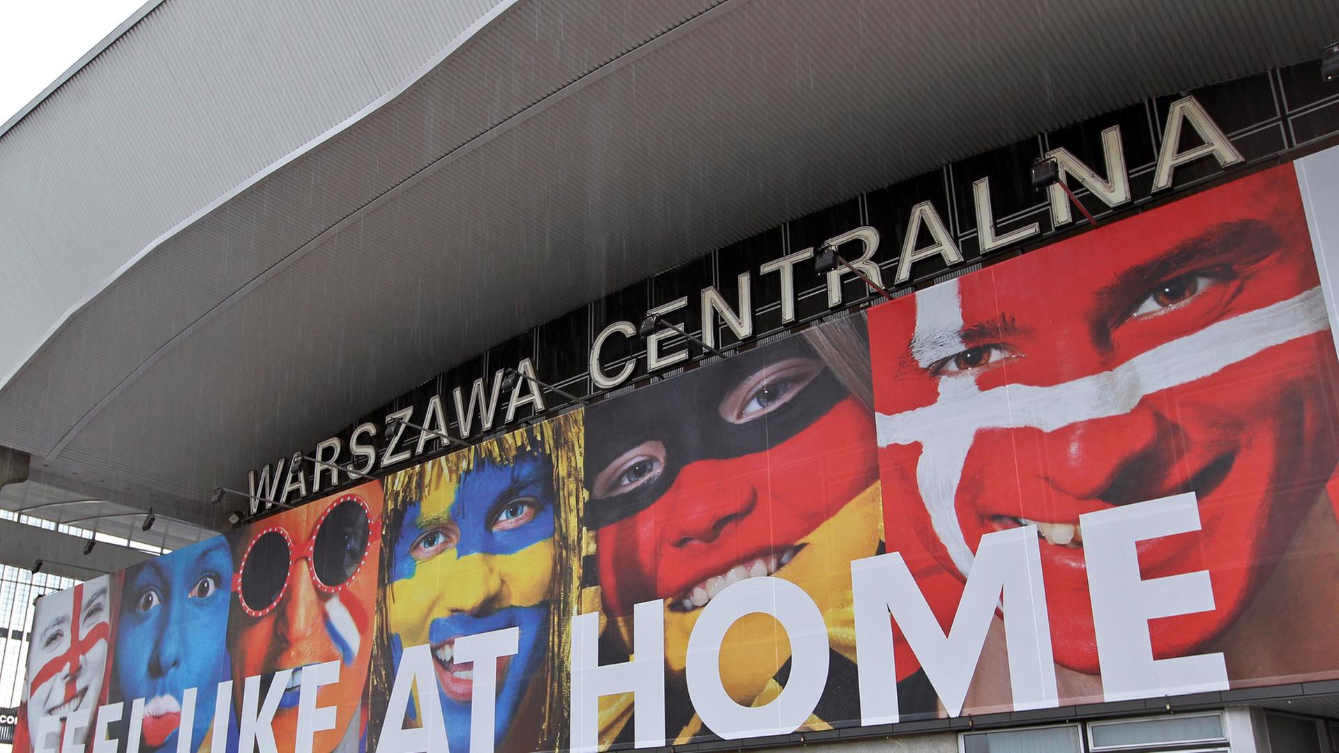 Ein riesiges Poster am Hauptbahnhof von Warschau empfängt Fußballfans aus ganz Europa zur Europameisterschaft 2012. Das Plakat trägt den Schriftzug "Feel like at home" und zeigt Menschen, die ihre Gesichter mit den Nationalflaggen von Teilnehmerländern bemalt haben.