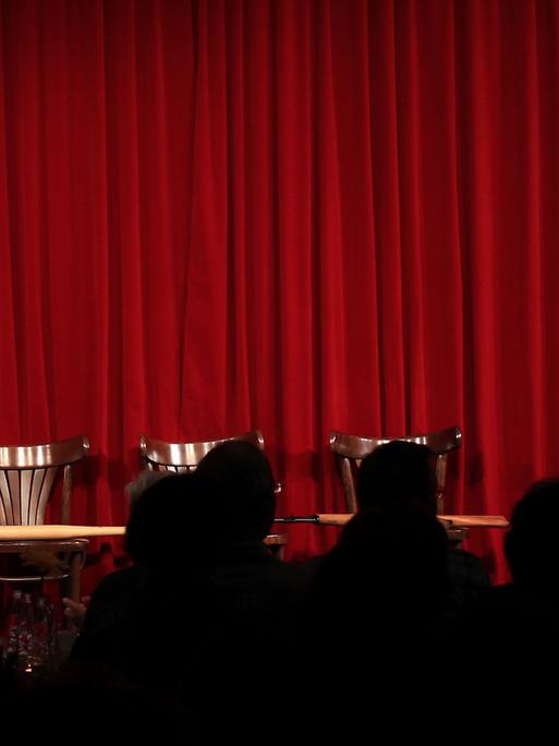 Zuschauer sitzen im Theater vor einer leeren Bühne mit geschlossenem roten Vorhang.