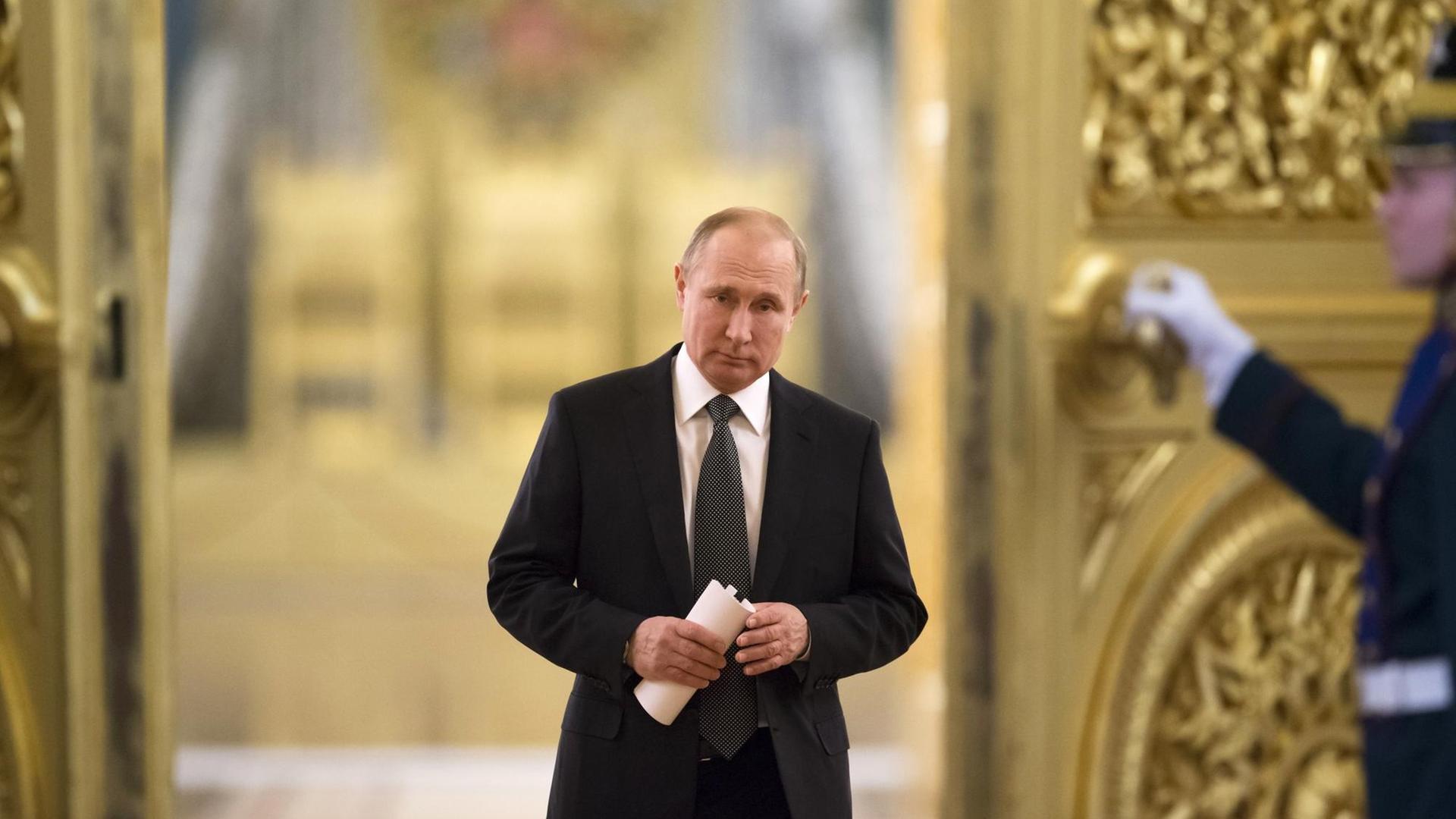 Das Bild zeigt Russlands Präsident Putin im Kreml. Er schreitet durch die Gänge und hält ein zusammengerolltes Stück Papier in seinen Händen.