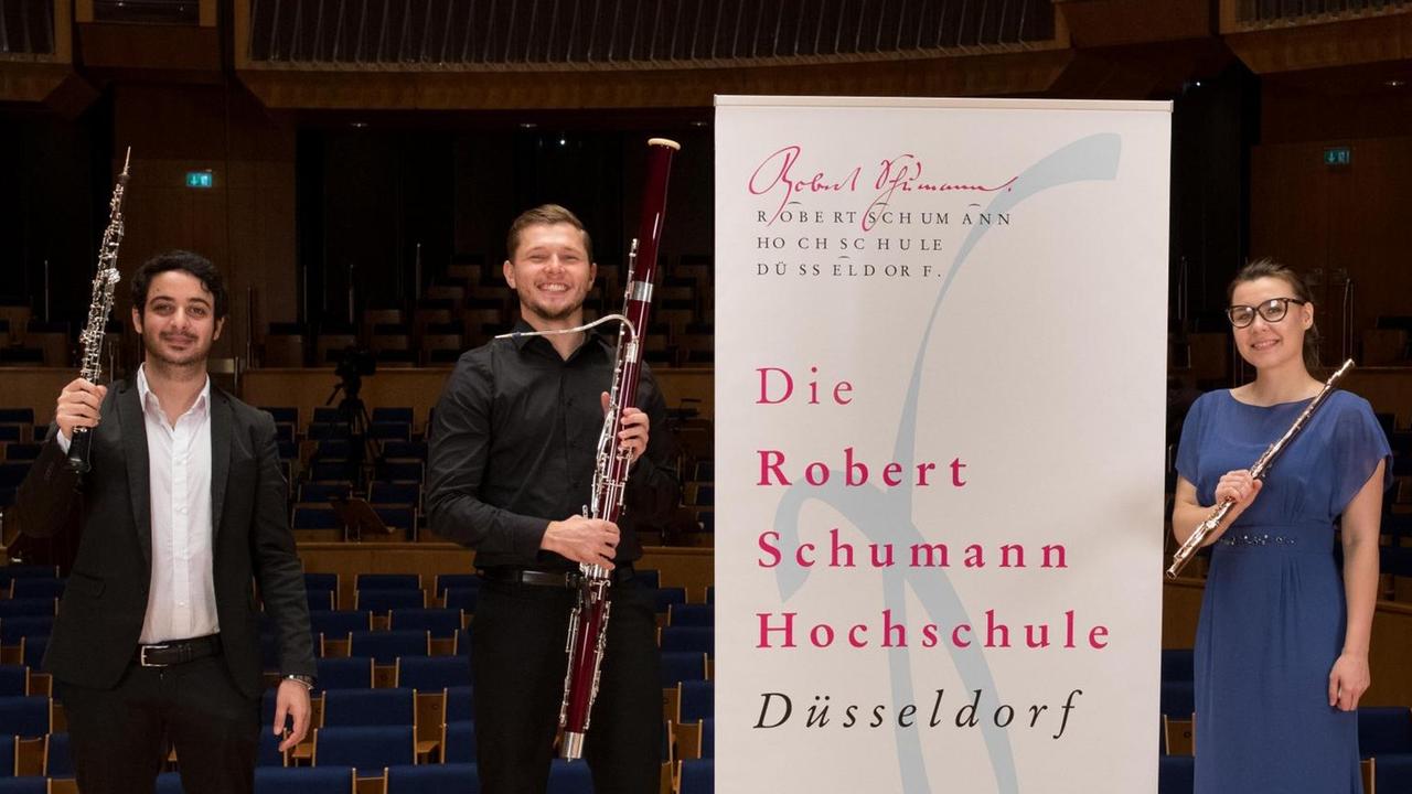 Ein Oboist, ein Fagottist und eine Flötistin stehen auf einer Bühne und posieren mit ihren Instrumenten für die Kamera. Zwischen ihnen prangt ein Banner der Robert Schumann Hochschule, Düsseldorf. Ein Oboist, ein Fagottist und eine Flötistin stehen auf einer Bühne und posieren mit ihren Instrumenten für die Kamera. Zwischen ihnen prangt ein Banner der Robert Schumann Hochschule, Düsseldorf.