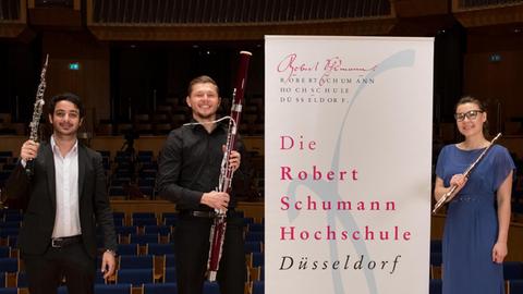 Ein Oboist, ein Fagottist und eine Flötistin stehen auf einer Bühne und posieren mit ihren Instrumenten für die Kamera. Zwischen ihnen prangt ein Banner der Robert Schumann Hochschule, Düsseldorf.