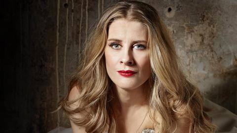Eine Frau trägt ihr langen blonden Haare offen. Sie sitzt in einem weißen Kleid vor einer Steinwand.