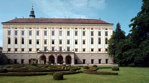 Blick auf das Schloss in Kroměříž, dahinter ist noch der Kirchturm der Schlosskapelle sichtbar. Von aussen ist es ein schlichtes rechteckiges Gebäude aus hellem Stein, mit rotem Dach, der Eingang ist ein kleiner Laubengang mit 4 Bögen. Davor befindet sich ein grüner Rasen mit kunstvoll geschnittenen Hecken