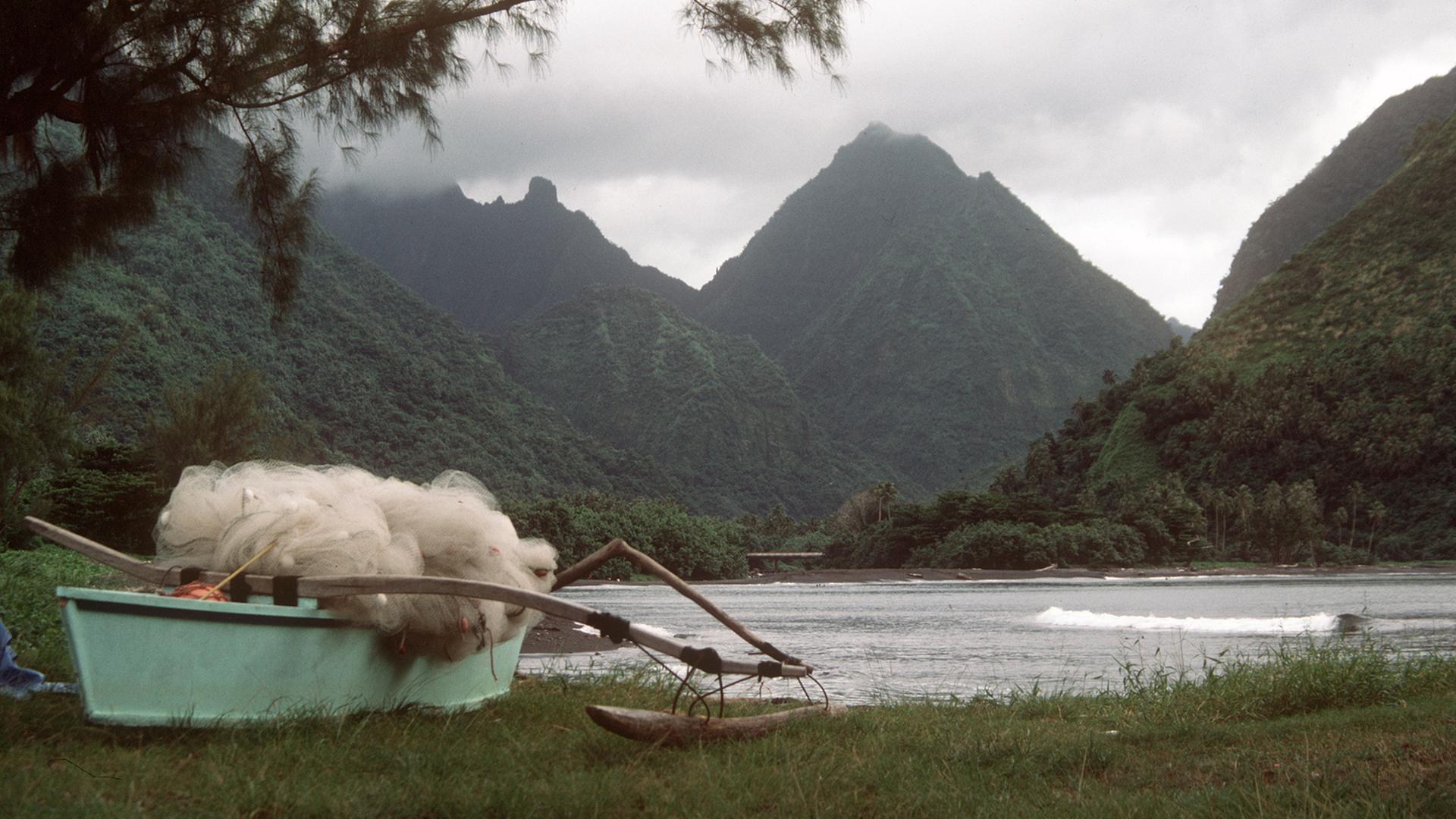 Eine Bucht in Tahiti mit dichter Vegetation und bizarr geformten Bergen, im Vordergrund ein typisches Auslegerboot mit Netzen von Fischern der einheimischen Bevölkerung. Aufnahme von 1985.