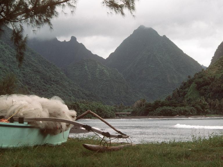 Eine Bucht in Tahiti mit dichter Vegetation und bizarr geformten Bergen, im Vordergrund ein typisches Auslegerboot mit Netzen von Fischern der einheimischen Bevölkerung. Aufnahme von 1985.