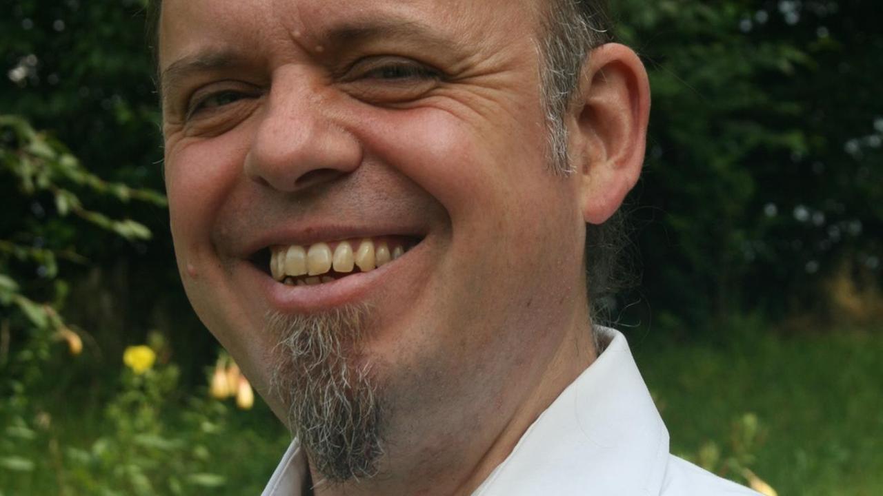 Porträt des Pfarrers und Übersetzers Matthias Binder, lächelnd, mit offenem weißen Hemd, im Garten