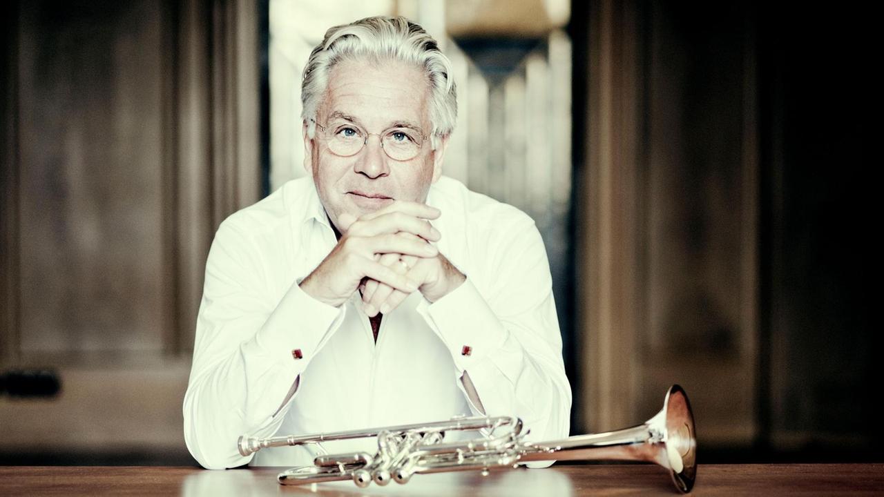Der Trompeter und Dirigent Håkan Hardenberger sitzt im weißen Hemd vor einem Tisch mit aufgestützten Armen, die Tropmete liegt quer vor ihm