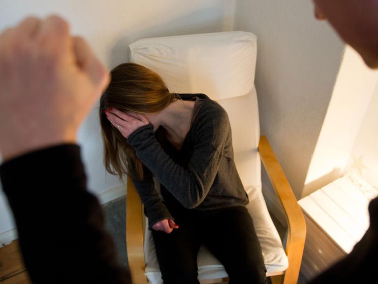 Ein Mann ballt seine Hand zur Faust, vor ihm eine Frau auf einem Sessel, die sich ängstlich die Hand vor ihr Gesicht hält.