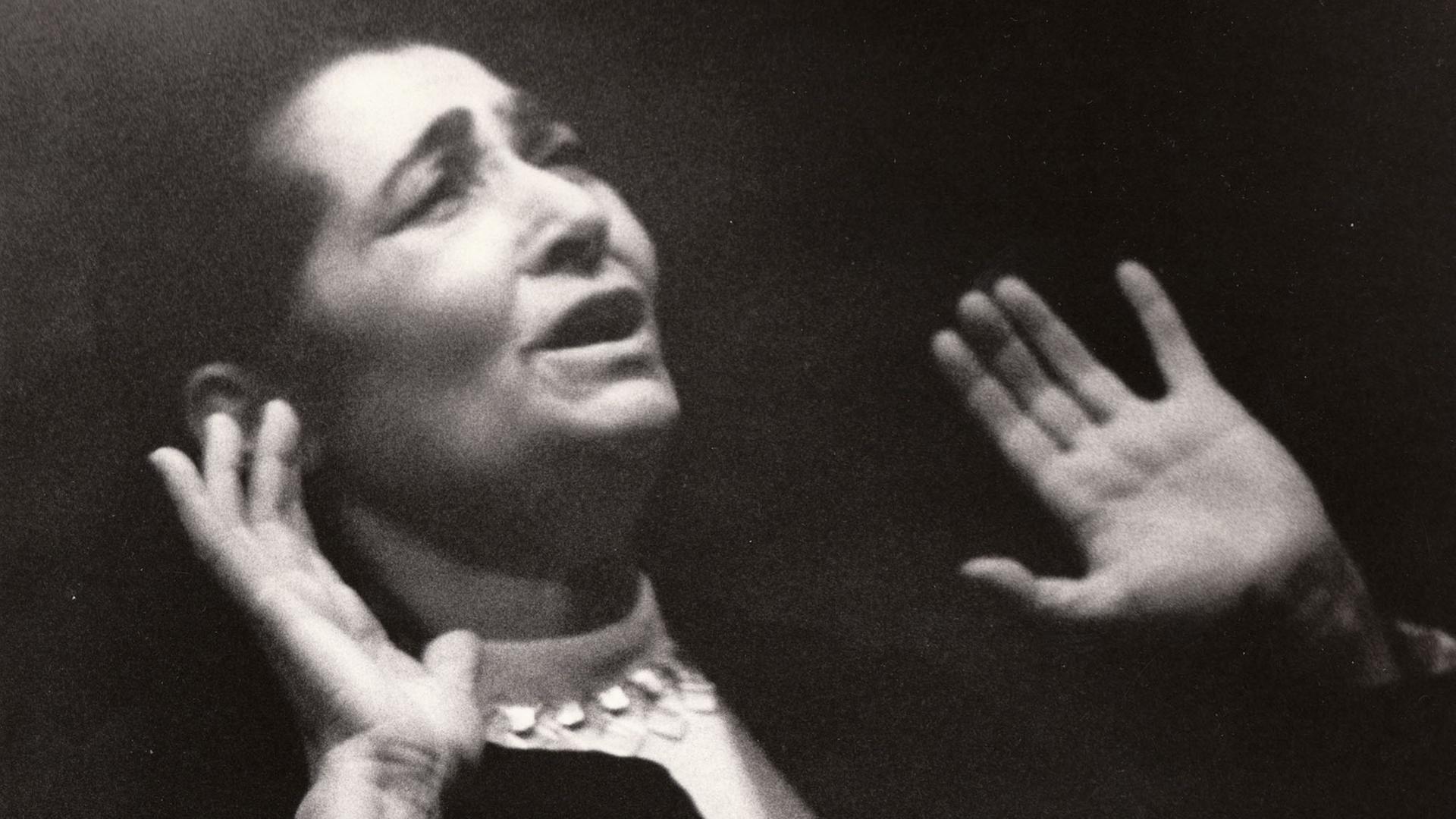 Schwarz-weiß-Aufnahme einer Frau, die mit dramatischem Gesichtsausdruck und nach oben gestreckten Händen singt.