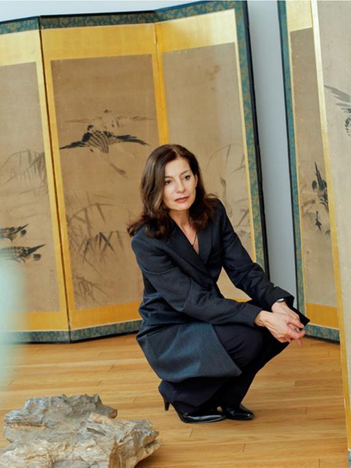 Die Schriftstellerin Ursula Krechel hockt gedankenversunken in einer Ausstellung. Auf Stellwänden sind Zeichnungen von Vögeln und Gräsern zu sehen.