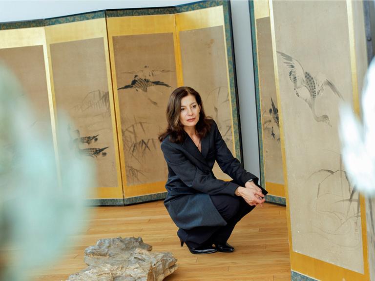 Die Schriftstellerin Ursula Krechel hockt gedankenversunken in einer Ausstellung. Auf Stellwänden sind Zeichnungen von Vögeln und Gräsern zu sehen.
