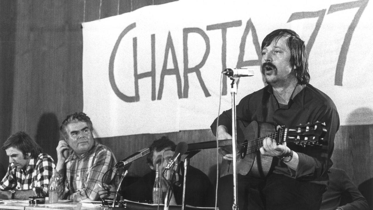Wolf Biermann singt in ein Mikrofon, hinter ihm auf einem Plakat prangt die Aufschrift "Charta 77".