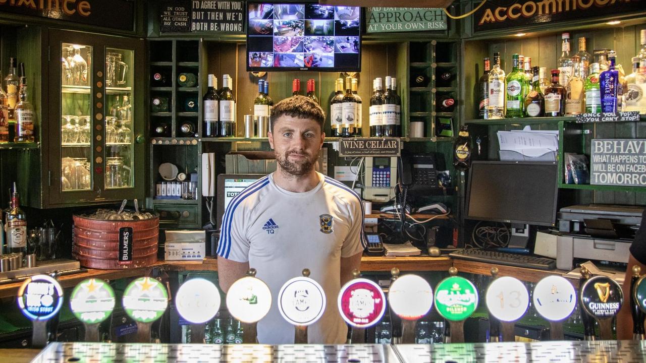 Tom Quinn im Traditionspub "Ferryman" in den Docks von Dublin. Er hat kurze Haare, ein Fußball-Trikot an und steht vor den Zapfhähnen.