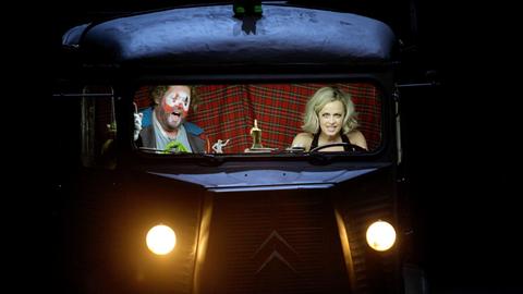 Eine Frau und ein Clown sitzen in einem Bus. Sie steuert das Fahrzeug und er singt,