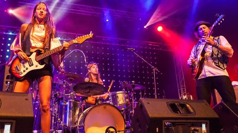 Eine Frau und zwei Männer stehen auf einer Bühne und spielen Gitarre bzw. Schlagzeug