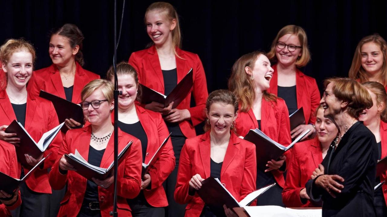 Die Mädchen des Chores stehen in ihrerer roten Konzertkleidung auf der Bühne und lachen einander zu.