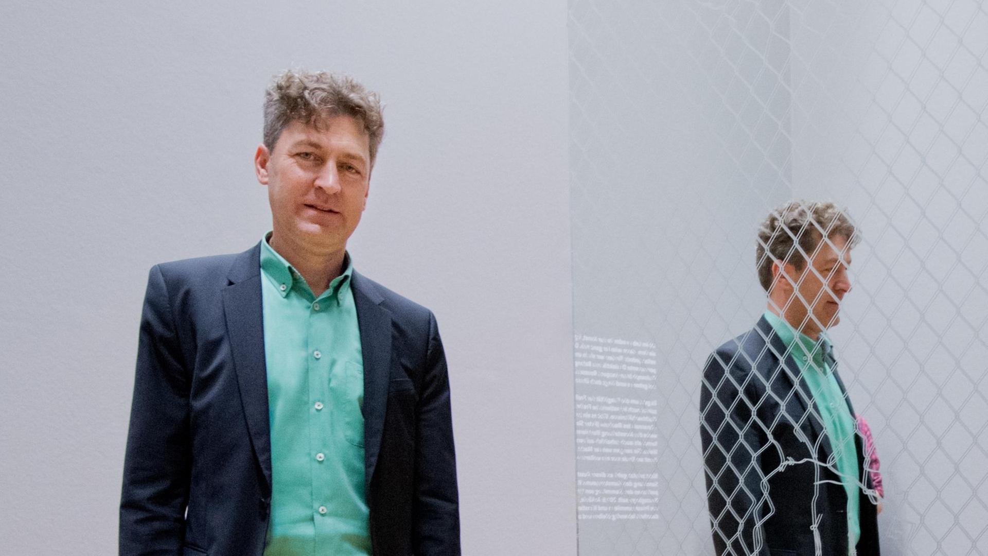 Ralf Beil steht vor einem grauen Hintergrund, er schaut in die Kamera, in einem Spiegel ist er hinter Maschendrahtzaun zu sehen.