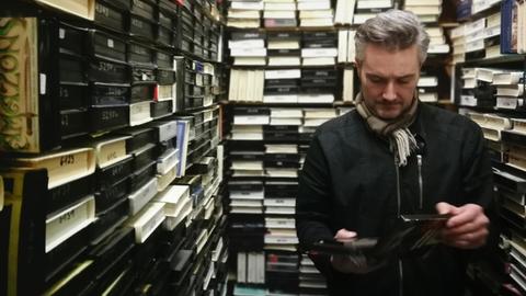 Ein Mann steht zwischen Regalen voller Videokassetten.