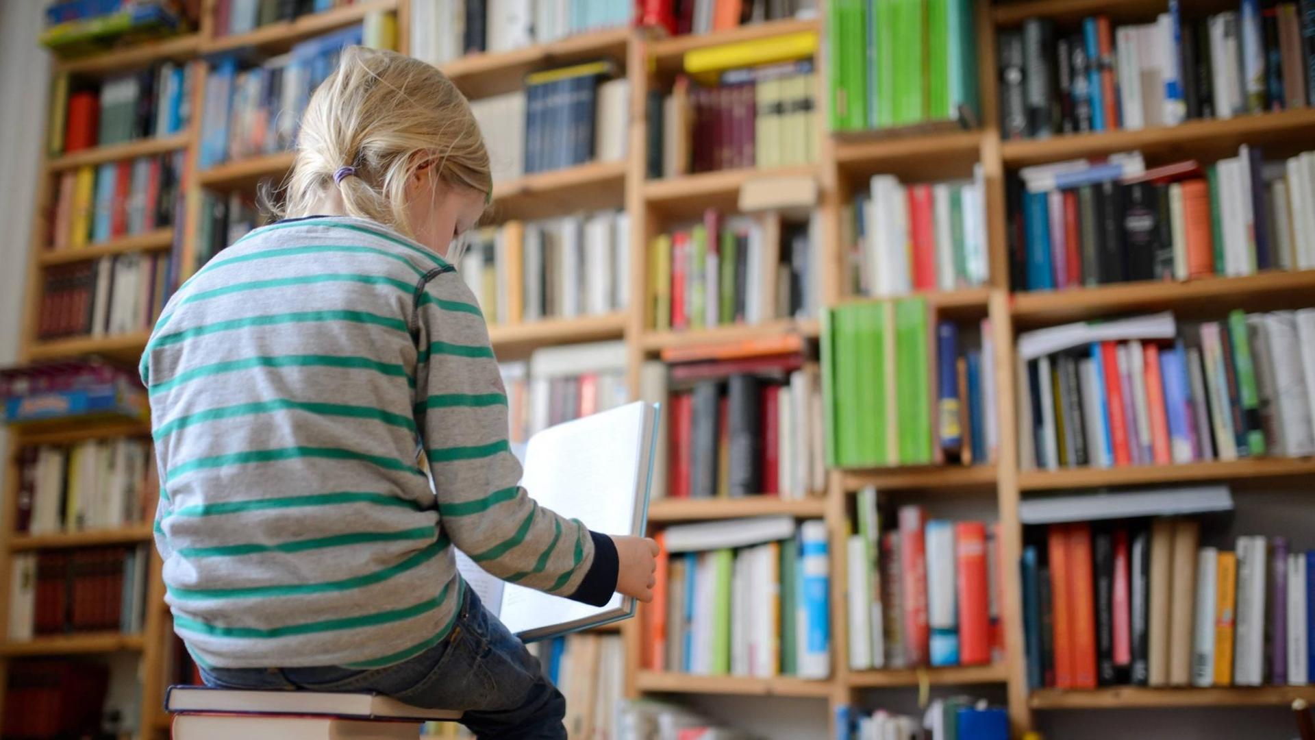Ein Kind sitzt auf einem Buchstapel vor einem Bücherregal und liest ein Buch.