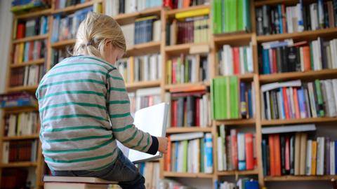 Ein Kind sitzt auf einem Buchstapel vor einem Bücherregal und liest ein Buch.