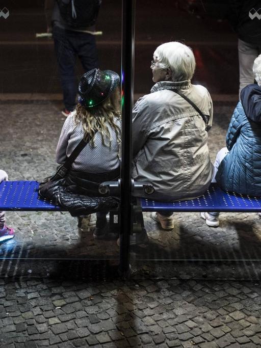 Drei Kinder und drei aeltere Menschen sitzen an einer Bushaltestelle in Berlin