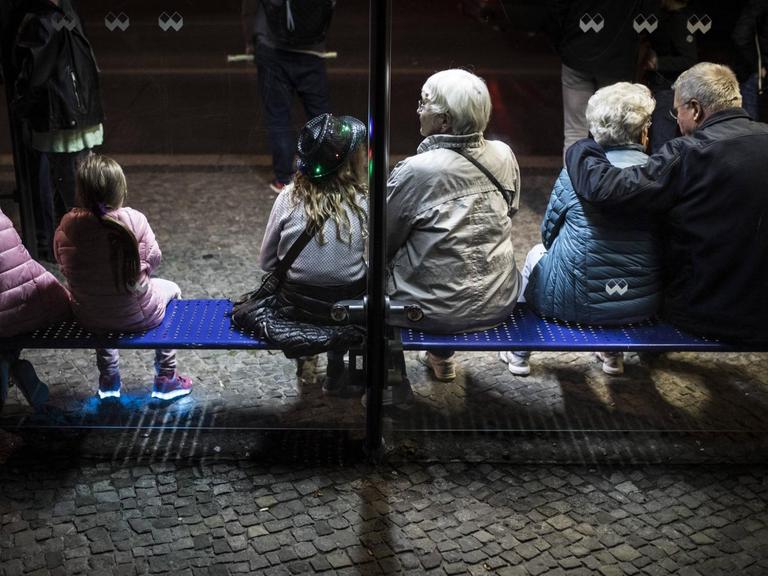 Drei Kinder und drei aeltere Menschen sitzen an einer Bushaltestelle in Berlin