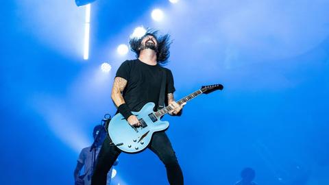 Dave Grohl spielt bei einem Konzert mit den Foo Fighters auf der Bühne Gitarre und wirft seinen Kopf mit den langen Haaren nach hinten.