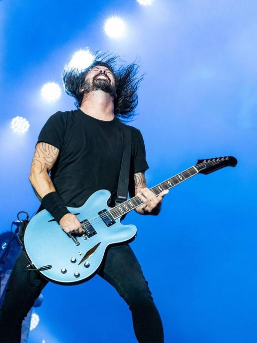 Dave Grohl spielt bei einem Konzert mit den Foo Fighters auf der Bühne Gitarre und wirft seinen Kopf mit den langen Haaren nach hinten.
