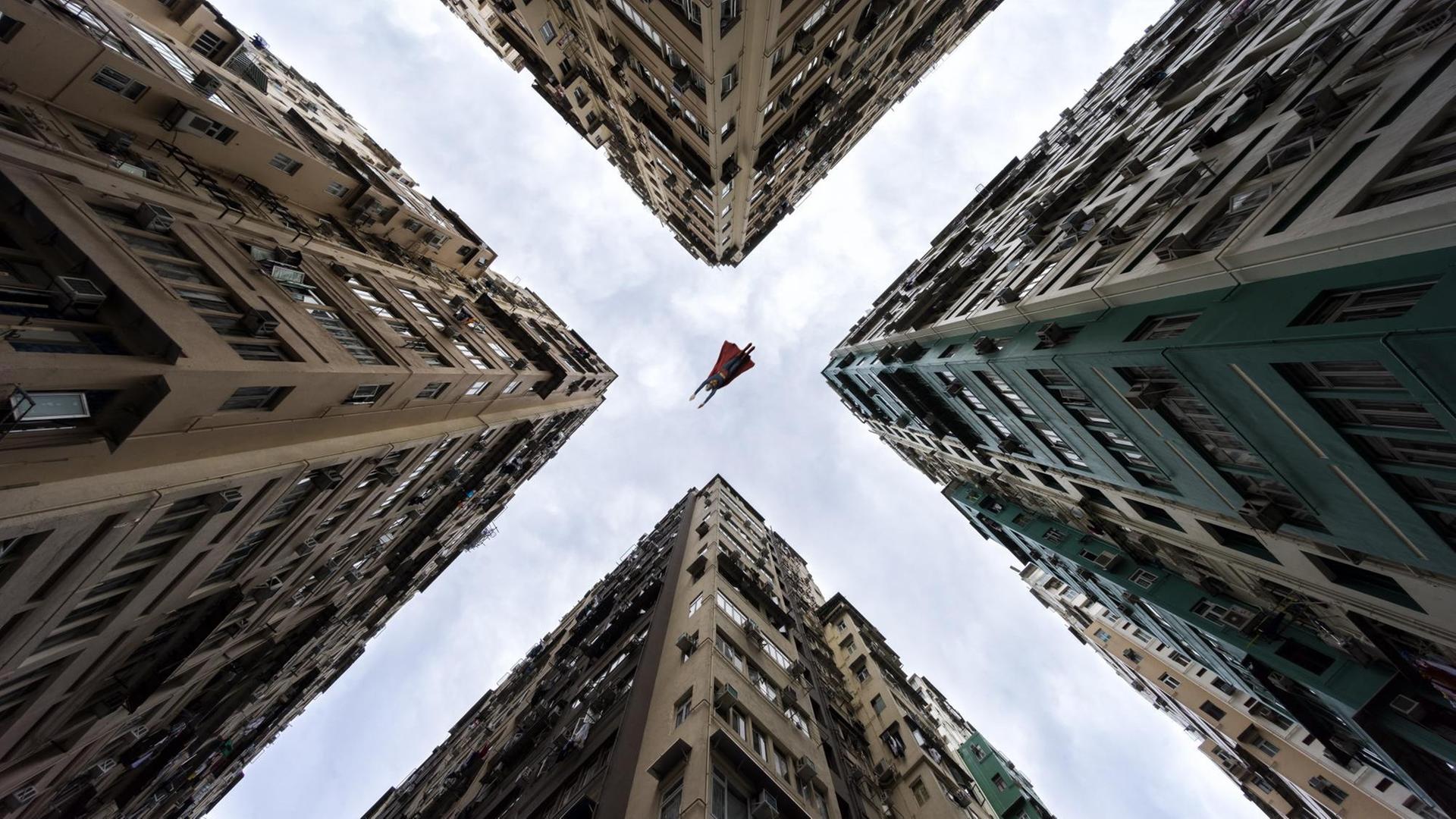 Aus der Froschperspektive sieht man Superman zwischen Häuserschluchten hindurch fliegen.